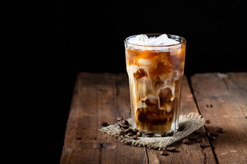 Ice coffee in a tall glass with cream poured over and coffee beans on a old rustic wooden table. Cold summer drink on a dark wooden background with copy space