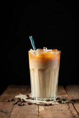 Ice coffee in a tall glass with cream poured over and coffee beans on a old rustic wooden table. Cold summer drink on a dark wooden background with copy space