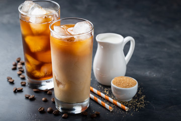 Ice coffee in a tall glass with cream poured over, brown sugar and coffee beans. Cold summer drink on a dark background. With copy space