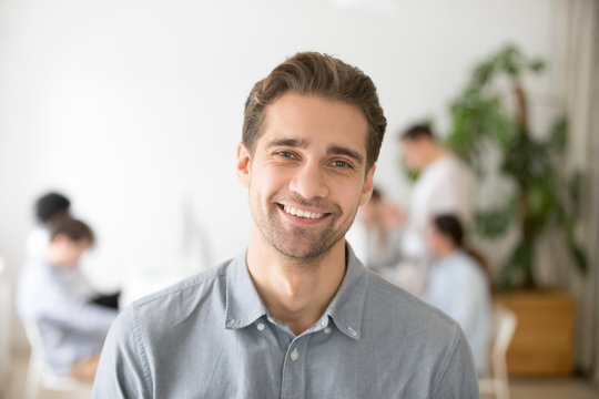 Portrait of casual smiling Caucasian male worker laughing looking at camera, positive employee posing for company business catalogue with colleagues at background, having photo shoot in office