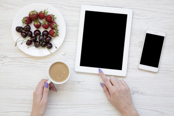 Feminine workspace with female hands, smartphone, tablet, berries and latte on white wooden background, top view. Flat lay. From above.