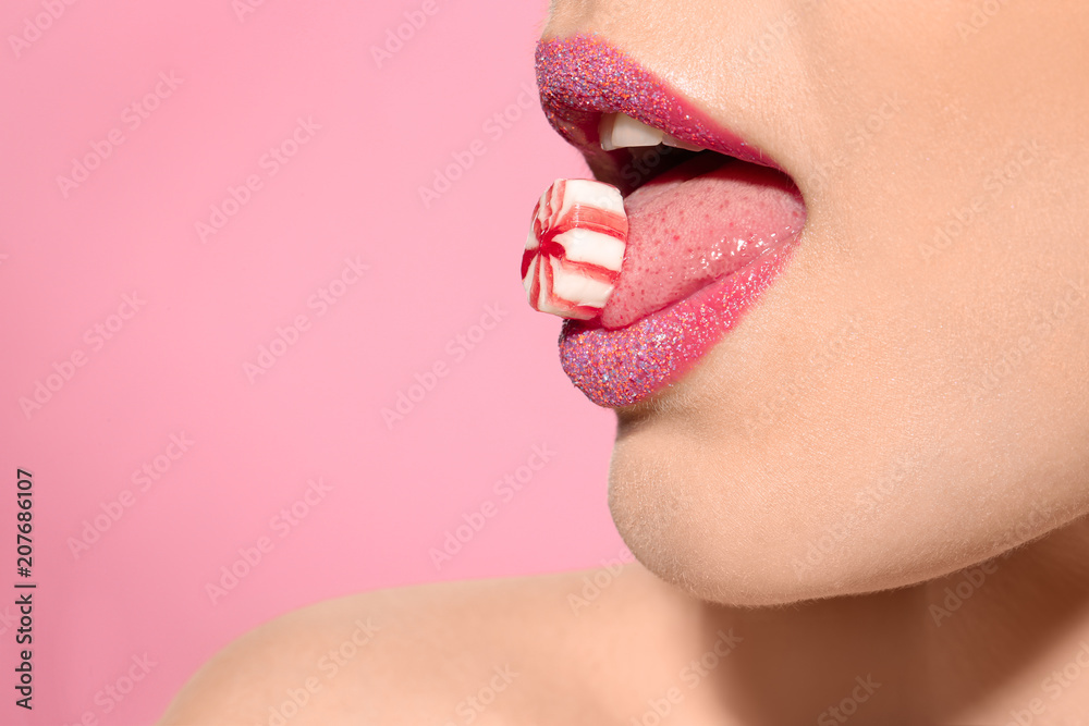 Poster Beautiful young model with creative makeup holding candy in lips on color background