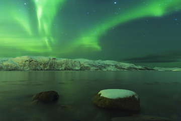 Northern lights Aurora borealis at the lakeside with rocks of a fjord during low tide in a snowy winter landscape with mountains