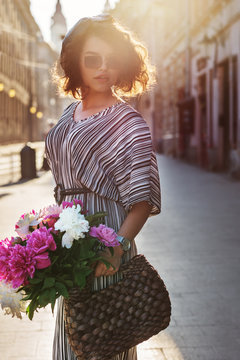 Outdoor portrait of young beautiful stylish curly girl wearing striped dress,  beret, rectangle sunglasses, holding straw bag with peonies, posing in street. Sunny day light. Summer fashion concept