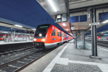 Passenger high speed train on the railway station at night in Europe. Urban landscape with modern commuter train on the railway platform with illumination. Intercity vehicle. Railroad travel