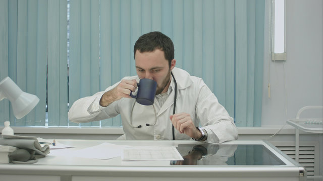 tired  from work, bearded doctor drink from cup and continue woking with documents and x-rays