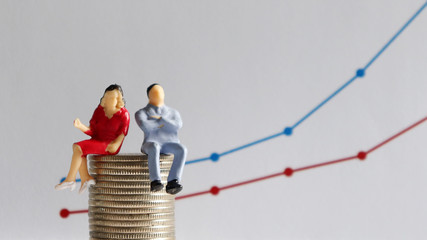 The concept of the wage gap between men and women in the workplace. A miniature man and a miniature woman sitting on a pile of coins in front of a linear graph.