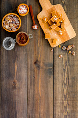 Cook caramel sauce. Melted caramel in glass jar, caramel cubes on cutting board, sugar and salt on dark wooden background top view copy space