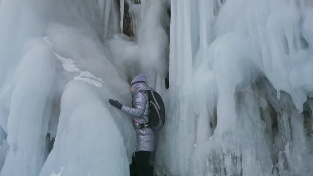 Travel of woman on ice of Lake Baikal. Trip to winter island. Girl is walking at foot of ice rocks. Traveler looks at beautiful ice grotto. Hiker wears sports glasses, silver jacket, backpack. Extreme