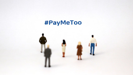 #PayMeToo as a new campaign to fill the wage gap between men and women. A miniature man and a miniature woman standing on a piece of paper with #PayMeToo on it.