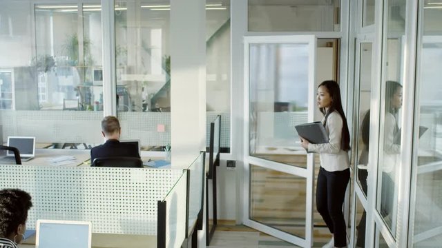 Tilt down of young Asian businesswoman walking with documents into modern open space office while employees working on computers at desks