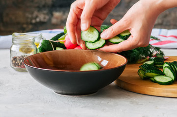 Healthy salad from fresh vegetables in making. Woman's hands putting slices of cucumber in a bowl. Cooking step by step concept.