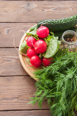 Fresh young vegetables for healthy salad. Radishes cucumbers dill spices and oil on a wooden plate. Wooden background.