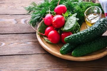 Fresh young vegetables for healthy salad. Radishes cucumbers dill spices and oil on a wooden plate. Wooden background.