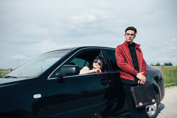 Pretty girl sitting behind the wheel of a black car on the background of a blue sky and a field next to a high handsome boy, in whose hands a business bag