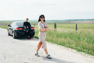 Couple of young girl and boy holding talks near black car on background of blue sky and field