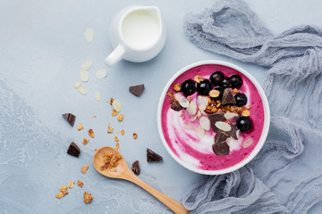 Pink black currant berries, banana smoothie bowl with granola, flakes from almonds and chocolate on light gray concrete background. Selective focus. Top view. Copy space.