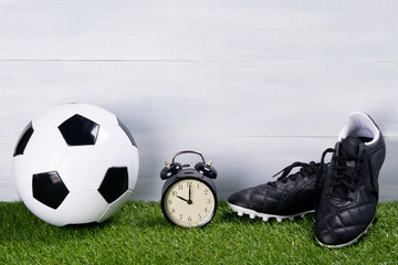 soccer ball, black boots and alarm clock, stand on the grass, on a gray background