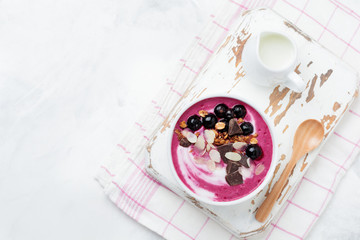 Pink black currant berries, banana smoothie bowl with granola, flakes from almonds and chocolate on light gray concrete background. Selective focus. Top view. Copy space.