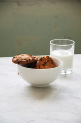 Homemade chocolate chip cookies in a bowl and glass of fresh milk on a white stone background.