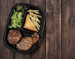 Grilled pork with french fries and salad in a plastic box on wood background