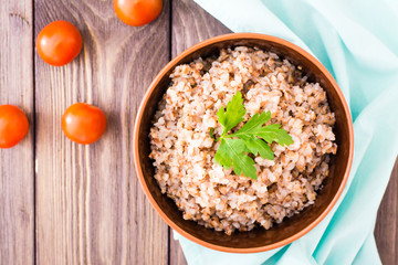 Cooked buckwheat in a bowl with a leaf of parsley and cherry tomatoes on a wooden table. Top view