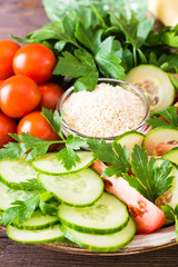 Fresh slices of cucumbers, tomatoes, sesame seeds in a bowl and parsley leaves on a plate