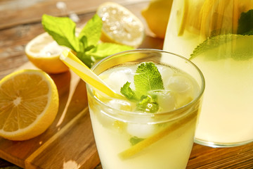 Lemonade pitcher and two glasses of cold refreshing citrus beverage with ice, lemon slices, mint leaves & straws on brown grunged wooden table, country side foliage background. Close up, copy space.