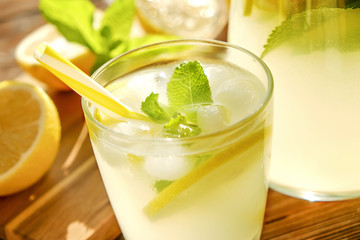 Lemonade pitcher and two glasses of cold refreshing citrus beverage with ice, lemon slices, mint leaves & straws on brown grunged wooden table, country side foliage background. Close up, copy space.