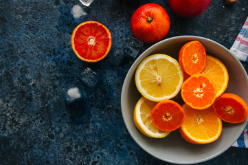 Mix of fresh ripe citrus fruits as blood oranges, mandarines, lemons  with ice cubes in a bowl on a blue stone background.