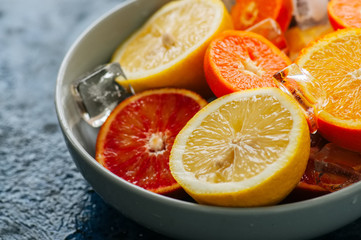 Mix of fresh ripe citrus fruits as blood oranges, mandarines, lemons  with ice cubes in a bowl on a blue stone background.