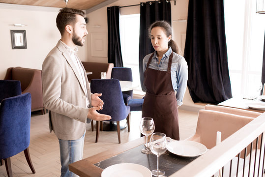 Displeased handsome restaurant manager with beard reporting young waitress in apron and gesturing while explaining her mistakes in modern establishment