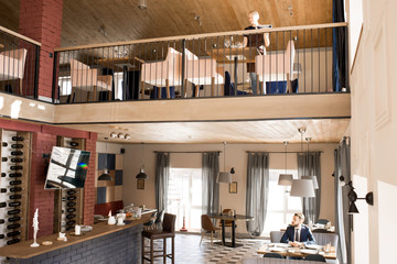 Serious pensive businessman sitting at table and looking around while waiting for order, young waitress setting table on the second floor