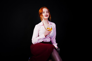 Gorgeous red haired girl in pink blouse and red skirt with glass of wine at hand isolated on black.