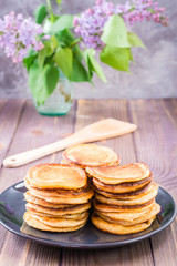 A pile of pancakes on a plate and a bouquet of lilac in a vase on a wooden table