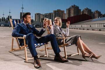 young happy business people relaxing on sunbeds and clinking glasses with coffee to go on parking