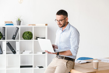 Image of businesslike man wearing white shirt sitting on table in office, and using silver laptop...