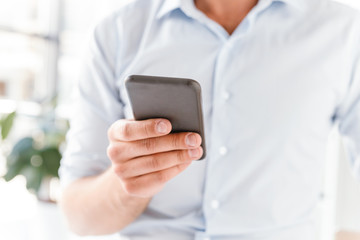 Cropped image closeup of european man in formal wear working in bright office room, and holding black smartphone in hand