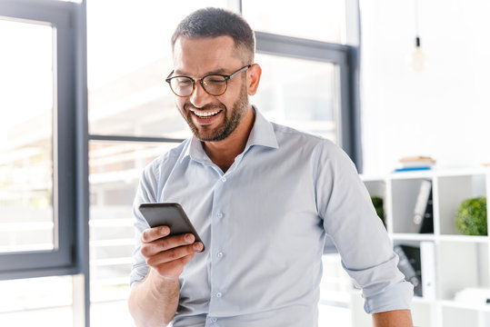 Image closeup of smiling employer guy in white shirt standing in office room near big window, and using smartphone for work