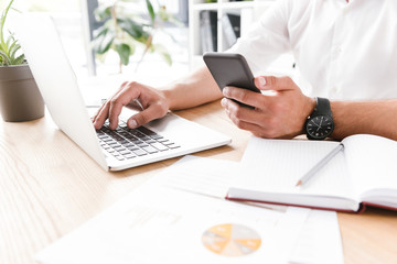 Cropped image of businesslike man in white shirt sitting at table and using smartphone with silver laptop, while working in bright office room