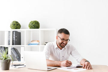 Photo of pleased businessman 30s in white shirt sitting at table and writing down information at notebook, while working with laptop in office