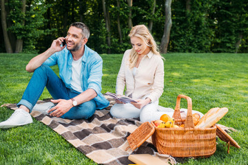 young woman reading book and smiling man talking by smartphone while sitting together on plaid at picnic in park