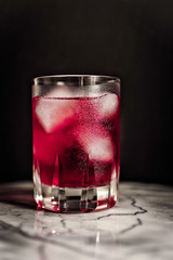 portrait of tumbler glass with red cold cocktail and ice cubes on marble table with black background