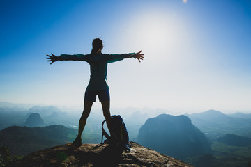 Successful cheering young woman hiker outstretched arms on sunrise mountain peak cliff edge