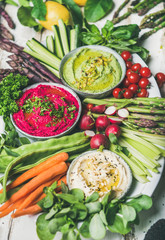 Healthy raw summer vegan snack plate. Chickpea, beetroot, spinach hummus dips with fresh vegetables and greens on white background, close-up. Clean eating, dieting, vegan or vegetarian party food