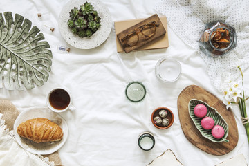 Creative flat lay of workspace in bed with croissant, cup of coffee, pastel pink macaroons, woman accessories on white bedsheets background. Flat lay, top view blogger influencer home office concept.