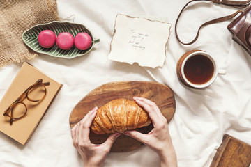 Creative flat lay with female hands on croissant, cup of coffee ,macaroons cakes, vintage card and glasses on white bedsheets background. Fashion and influencer blogging concept.