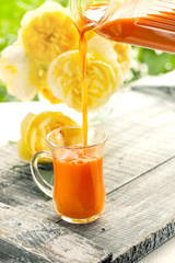 Carrot juice is poured from a jug into a glass Cup on a background of roses
