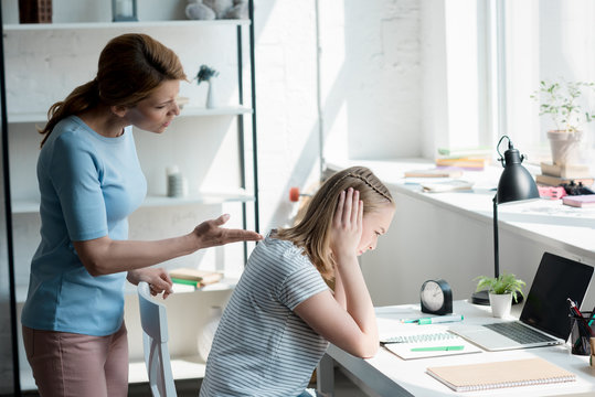 Depressed Teen Daughter Sitting At Work Desk At Home While Her Mother Yelling At Her