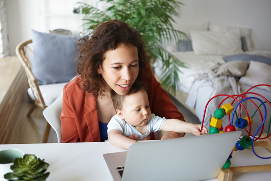 Picture of happy successful young businesswoman combining career and motherhood, working remotely using generic portable computer at home with her toddler son sitting on her lap and playing with toy